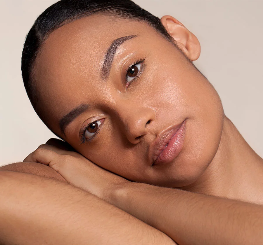 Close-up portrait of a young woman with dark brown skin and sleek, pulled-back hair. Her gaze is directed downward, and her face, with its subtle shine and dark circles beneath the eyes, exudes a sense of alluring mystery. The soft lighting and minimalist background create a captivating and intimate portrait, emphasizing her natural beauty and conveying a sense of serenity and introspection. - Bellafil in Thiensville, WI
