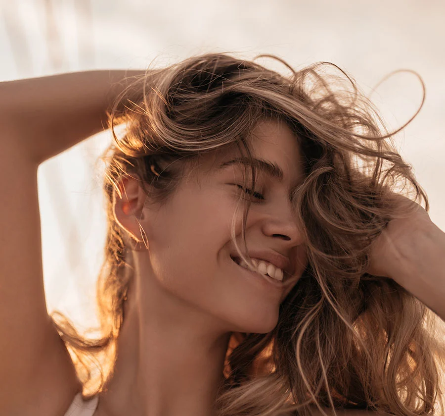 Close-up portrait of a woman with long, light brown hair enjoying a moment of pure joy. Her hair is tousled by the sunlight, and her eyes are closed as she smiles radiantly. The soft, warm light and the blurred background create a captivating and intimate portrait, capturing a moment of pure happiness and carefree bliss. - Broad Band Light Therapy in Thiensville, WI