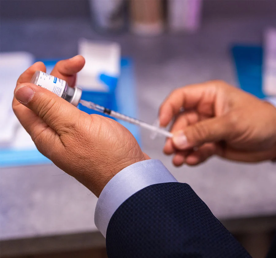 Close-up of gloved hands carefully preparing a medical injection. A vial containing a white liquid is held securely, while a syringe is being attached. The blurred background suggests a clinical setting, with medical supplies visible. The image highlights the precise and careful nature of medical procedures. - Dermal Fillers in Thiensville, WI