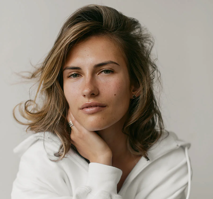 Close-up portrait of a woman with shoulder-length, light brown hair and a calm, inviting gaze. She wears a simple white hoodie and rests one hand gently on her neck, creating a sense of quiet confidence. The soft lighting and neutral background highlight her natural beauty and create a serene and intimate portrait. - Halo Laser in Thiensville, WI