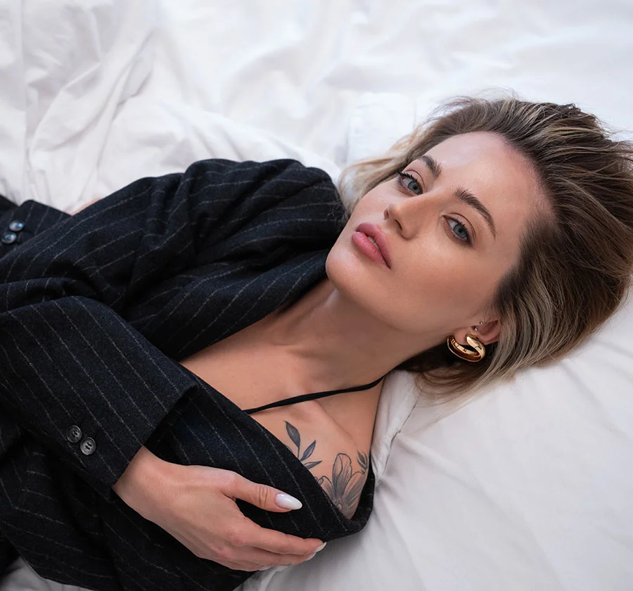 A young woman with shoulder-length light brown hair lies on a white bed, wearing a stylish gray blazer draped over her. Her neutral expression and subtle makeup, combined with the soft lighting and minimalist background, create a captivating and intimate portrait. The image highlights her natural beauty and effortless style. - Laser Tattoo Removal in Thiensville, WI