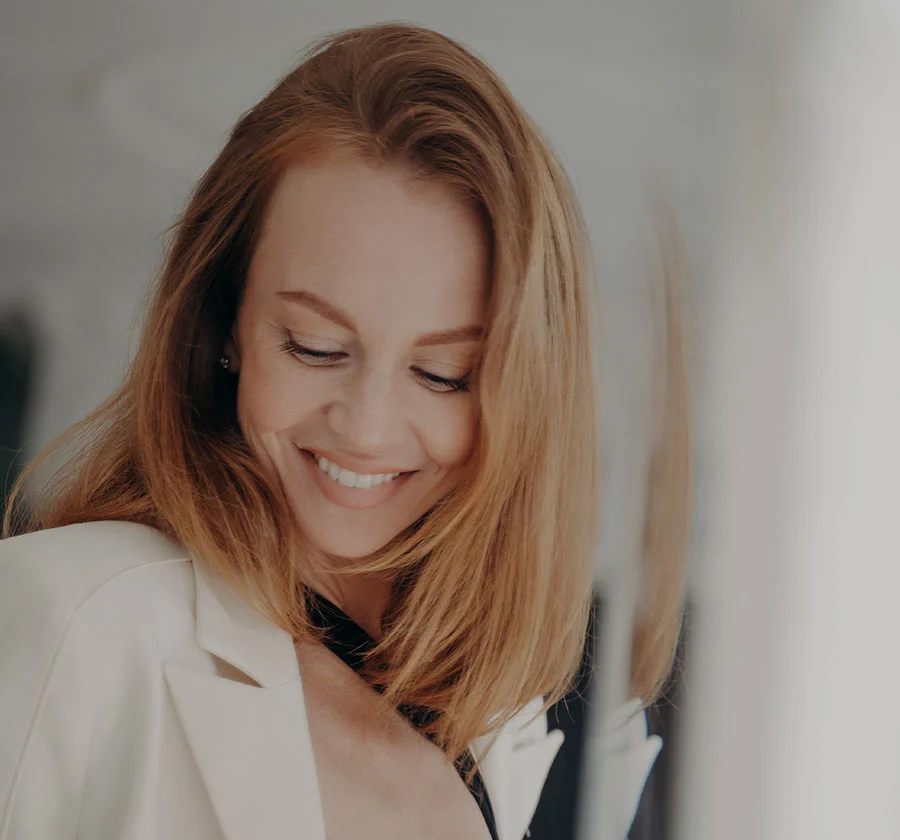 Close-up portrait of a woman with shoulder-length, wavy brown hair. She has a gentle smile and her eyes are closed, conveying a sense of peace and contentment. She wears a light beige blazer, and the soft, warm lighting creates a natural and inviting atmosphere. The image celebrates the beauty of a relaxed and confident woman. - O Shot and G shots in Thiensville, WI